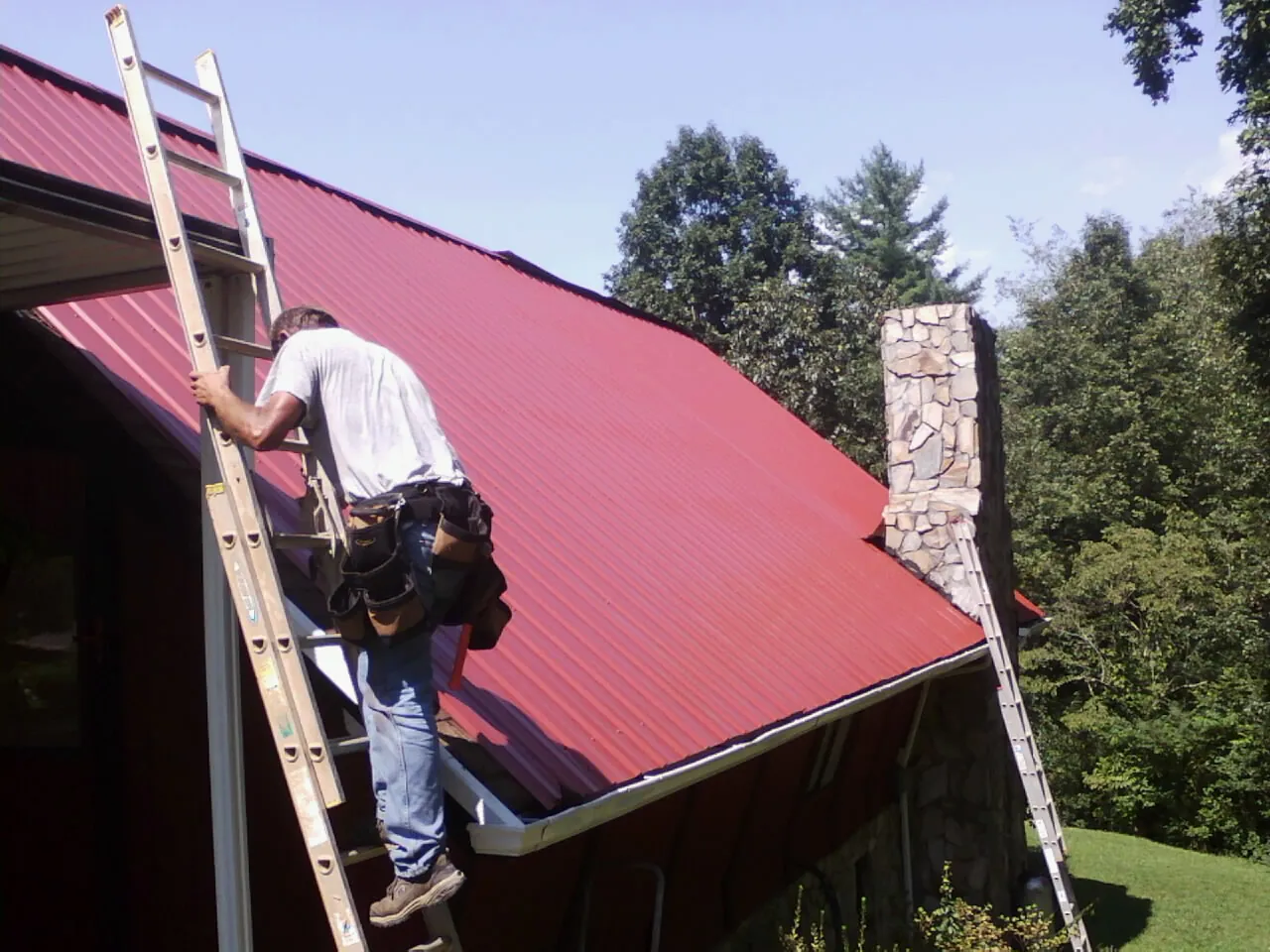 Roofer on a ladder beside a red metal roof and stone chimney — Soffit & Fascia Repair by Roofing Brothers in Mentcle